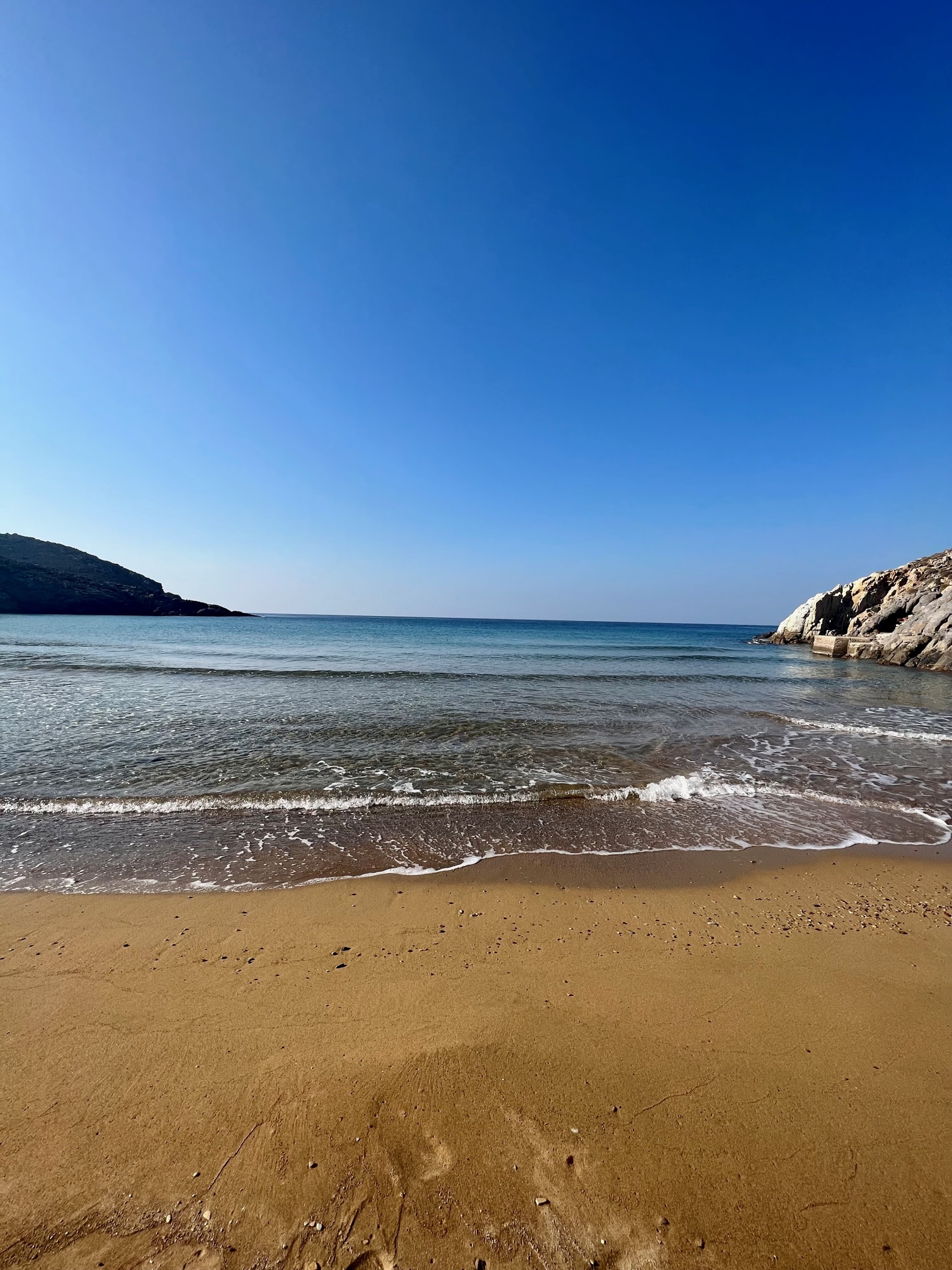 Sandy beach with gentle waves and rocky cliffs in Patmos island